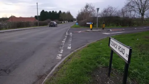 BBC A rural road bordered by grassy verges and leafless trees. A black car is driving away in the distance. On the right side of the image, a white street sign reads “TODWICK ROAD S26” near a junction with traffic islands and bollards.