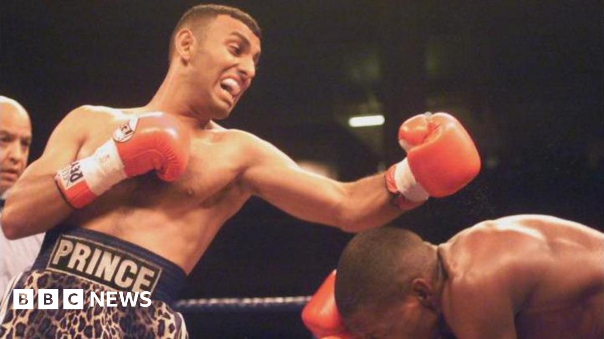 Prince Naseem Hamed punches with his opponent, who bows down to try to avoid the hit, with his left hand in a red boxing glove in the ring. He wears shorts in leopard print and the word Prince on his belt around the waist.