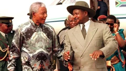AFP via Getty Images An old photo of Nelson Mandela talking to Yoweri Museveni, who wears one of his signature wide-brimmed hats and a suit