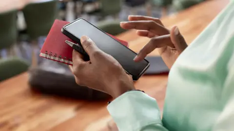 Getty Images Woman types on phone while holding a notebook and a pen