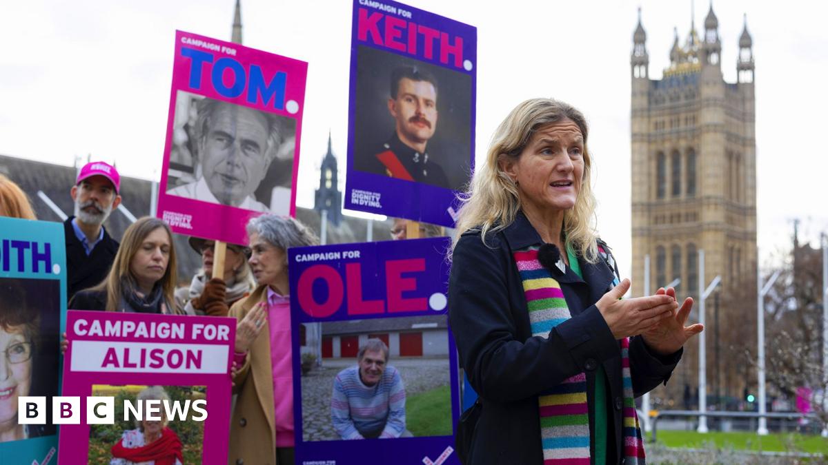 Labour MP for Spen Valley Kim Leadbetter wears a colourful stripey scarf while gesturing and speaking in front of parliament with campaigners in the background holding placards showing faces of people affected by the current law against assisted dying, in London in December.