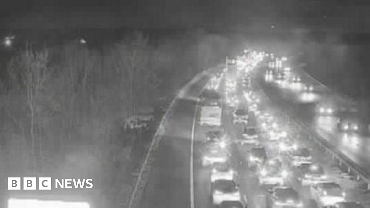 A traffic camera image above the M11 motorway. It is night time and the vehicles on both sides of the carriageway have their lights on. The carriageway closest to the camera has long queues of traffic.