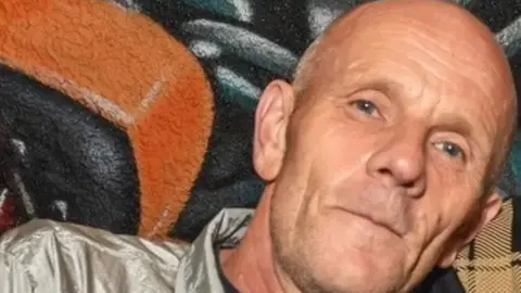 GoFundMe Paul Lumber looks at the camera. He is in his early 60s, white, bald and with light stubble. He is wearing a grey, shell-suit like top. 