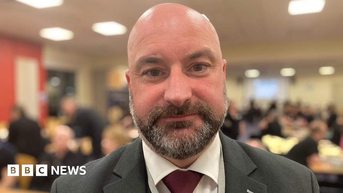 Head and shoulders shot of Marc Jones. He is wearing a grey suit and red tie and is looking directly at the camera. The background of the image is blurred but shows people sitting and standing at desks.