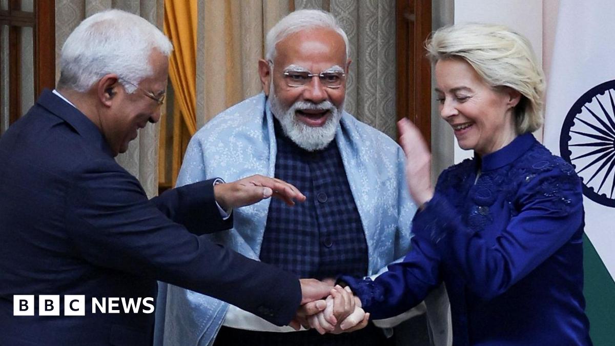 Three leaders are standing close together indoors, smiling and laughing during a friendly moment. The man in the centre, Indian Prime Minister Narendra Modi, with a white beard and glasses, wears a light shawl over a dark outfit and appears animated. On either side, European Council President António Luís Santos da Costa (left) in a dark suit and European Commission President Ursula von der Leyen (right) a deep blue jacket lean in, clasping hands in a gesture of warmth. A national flag and curtains are visible behind them