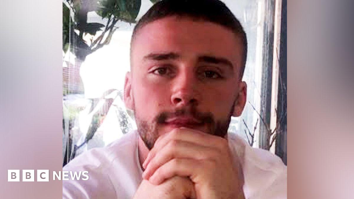 A young man with short, dark hair and stubble looks into the camera with his hands clasped in front of his chin. He is wearing a white T-shirt and is sitting in front of a glass pane.