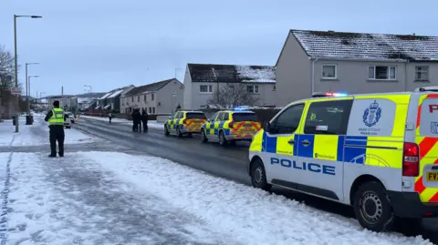 BBC A police van and an officer in a hi-vis vest over black clothing standing next to a snow path in Inverness.