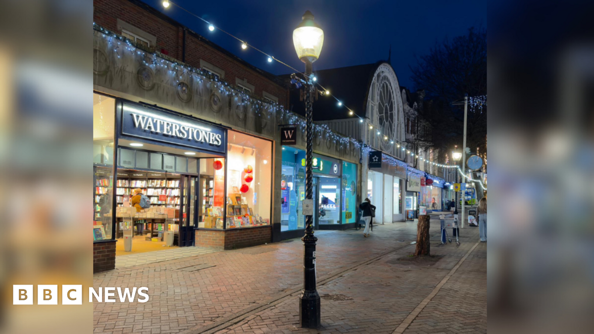The image shows a high street with lit up shops like Waterstones and Superdrug. In the centre of the image is a streetlight that has Christmas lights hanging from it. A white sheet has been stuck to the neck of the structure.