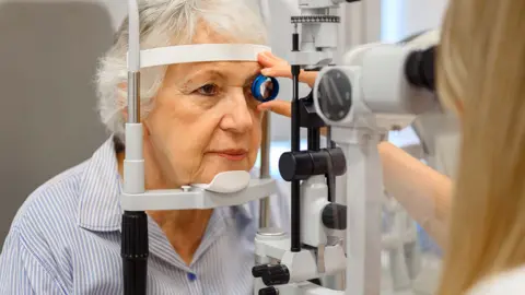 Getty Images An older lady having an eye test. She is looking into a machine while  an optician hold a lens to her left eye.