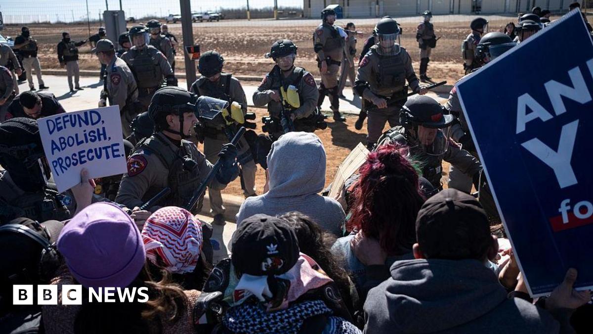 a group of protesters, holding anti-ICE signs, stand in front of a group of state troopers on a desert road