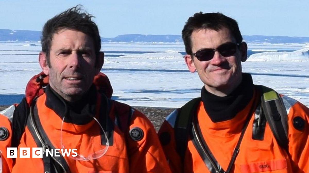 Prof Dave Roberts and Prof Colm O'Cofaigh in bright orange gear and looking at the camera. They are standing in front of an ice sheet. O'Cofaigh is wearing black sunglasses. They both have brown hair.
