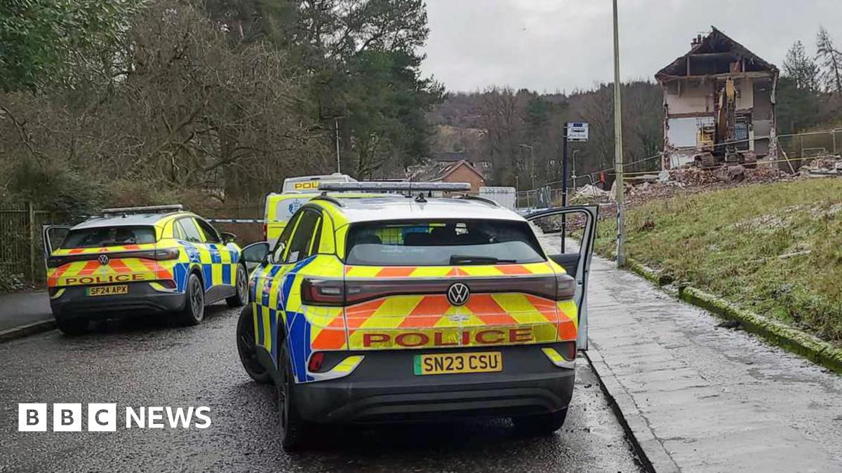 Two police vehicles parked on a residential road near a severely damaged house, with debris visible and trees lining the street