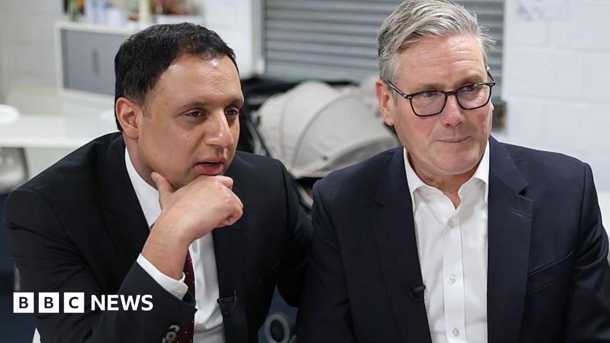 Anas Sarwar, in a dark blue suit and white shirt with a burgundy tie, sits with his chin resting on his hand. To his left, on the right of the shot, is Sir Keir Starmer in a blue suit and white open-necked shirt. Sir Keir is wearing thin, black plastic glasses. They are looking in the same direction off screen and appear to be listening. They are in a white, functional room with a pram behind them.