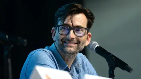 Getty Images Tennant wearing a blue button-up shirt speaking into a microphone at a panel discussion, with name placards visible in the foreground