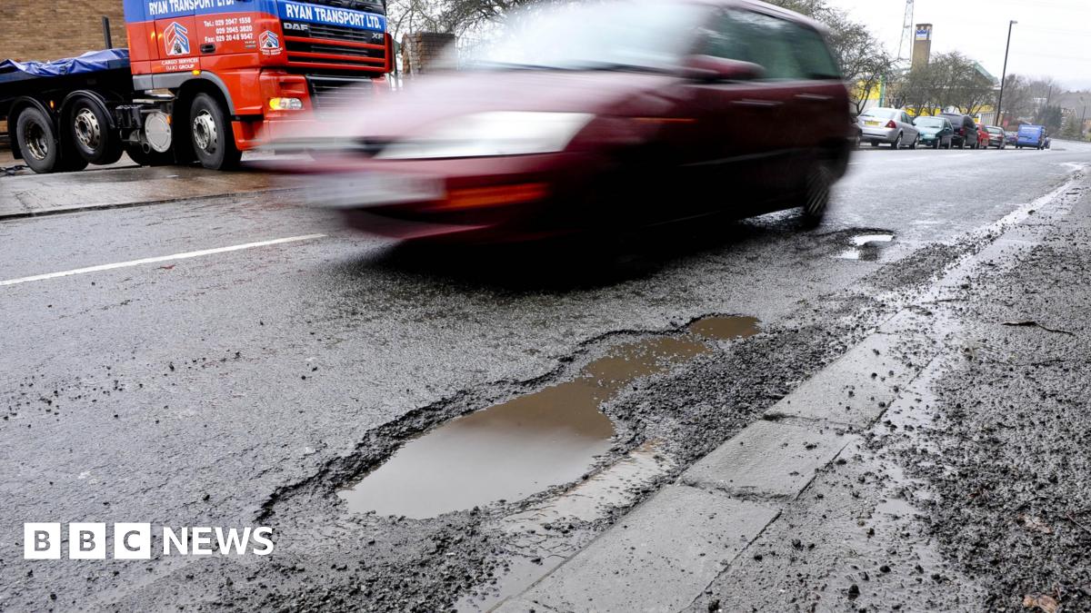 A car drives past a pothole on a road. Other vehicles can be seen on the road.