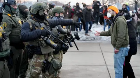 Reuters A federal agent wearing camouflage gestures towards a demonstrator wearing jeans and a green jumper and a face maskat a protest against the fatal shooting of Renee Nicole Good by a U.S. Immigration and Customs Enforcement (ICE) agent in Minneapolis, Minnesota