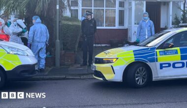 A semi-detached house with police cars parked outside. Three people in fill forensic suits are in view.