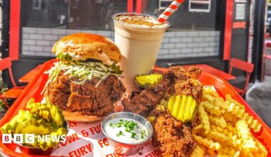 Photograph of Justina John, owner of Onja restaurant. She wears a black polo top which reads "Onja, taste of tanzania" on the yellow circle logo. Her hair is tied up into a black fishnet and she smiles at the camera.