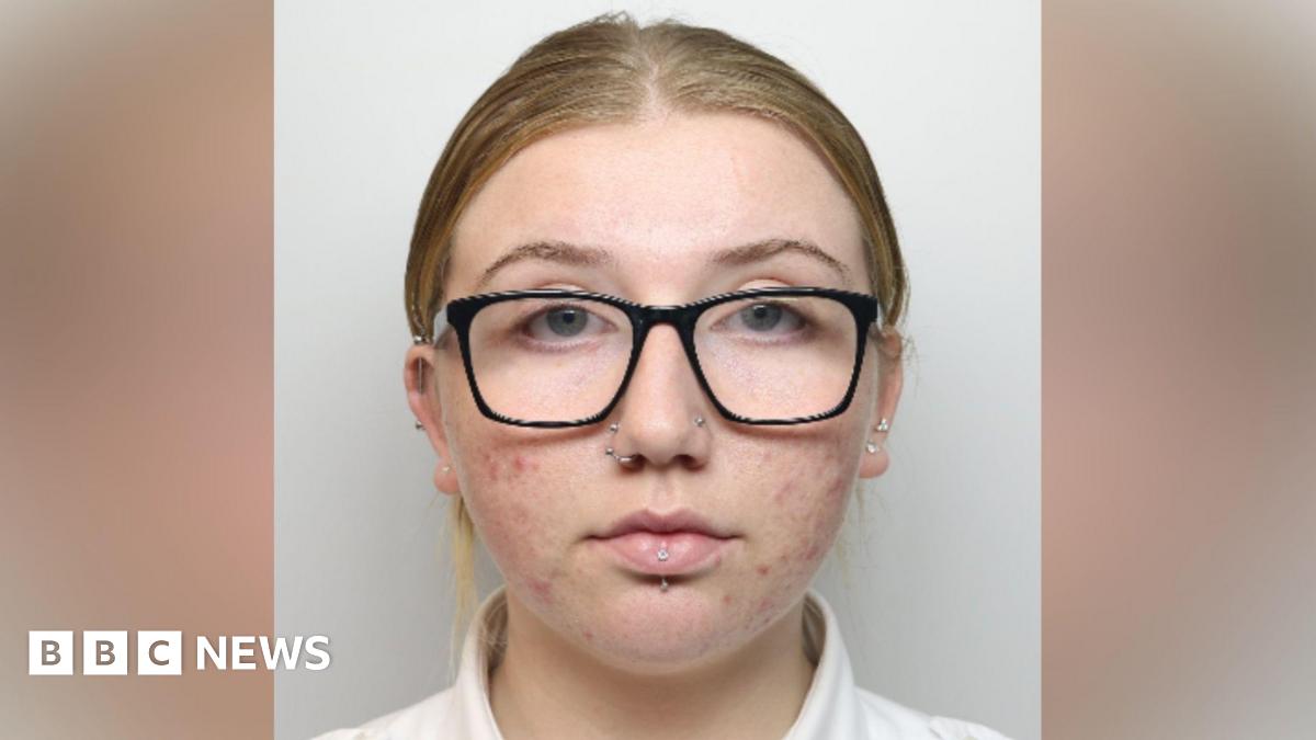 A woman with black-framed glasses, two nose piercings and a lip piercing looks at the camera.