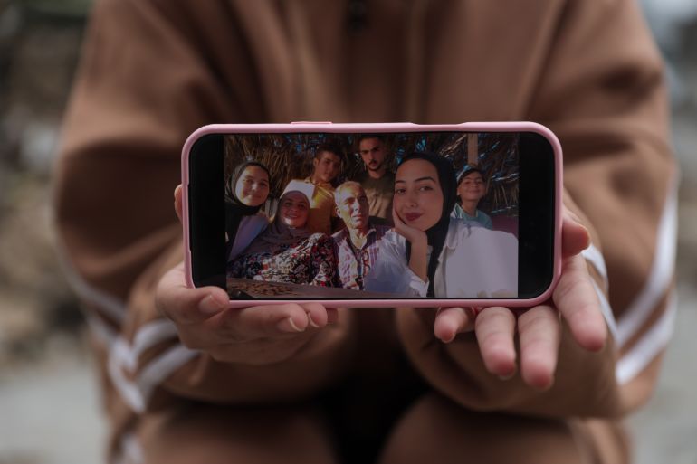 Batoul holds a photo on her phone showing her with her family, including her father, mother, and siblings Muhammad, Youssef, Tayma, and Habiba [Abdelhakim Abu Riash/Al Jazeera]