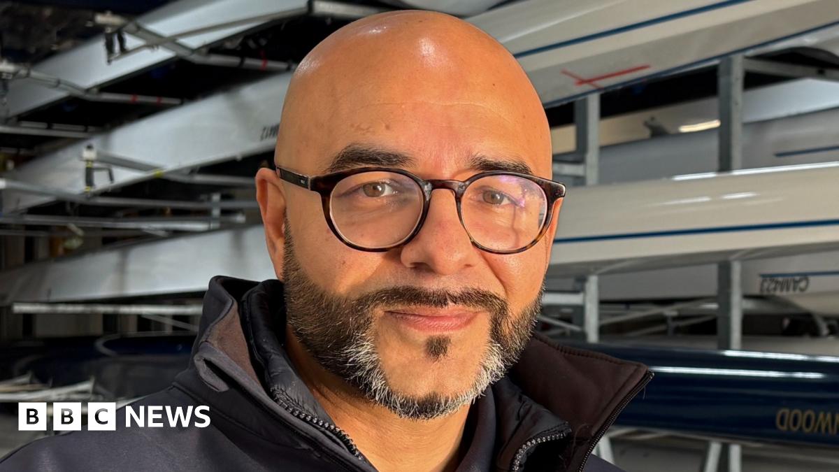 Omar Terywall, to the left of centre frame and looking directly out of the image. He is smiling slightly, has a dark, neatly trimmed beard, flecked with grey on his chin, and has on circular, black-framed glasses. He is bald. He is wearing a dark blue, collared top, with a white 'Cambridge Rowing' insignia on the right. Behind Omar are several sporting rowing boats mounted on racks inside a boathouse.