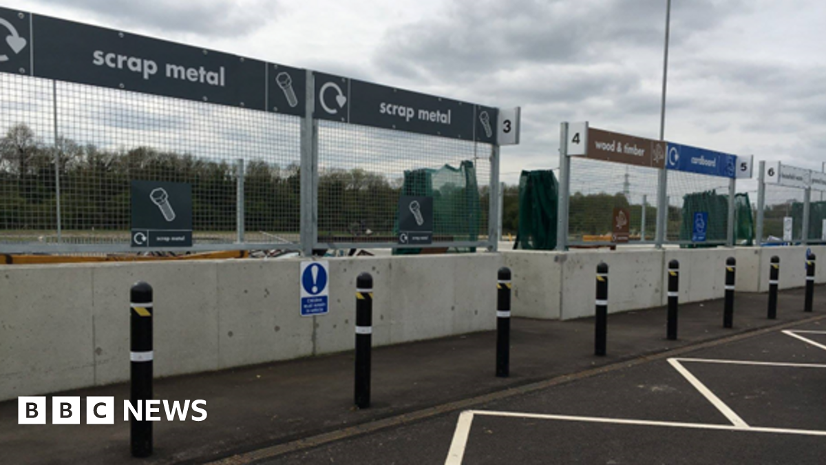 A household waste recycling centre with signs for scrap metal, wood and timber and cardboard. In front of the signs there are concrete barriers and a line of bollards.