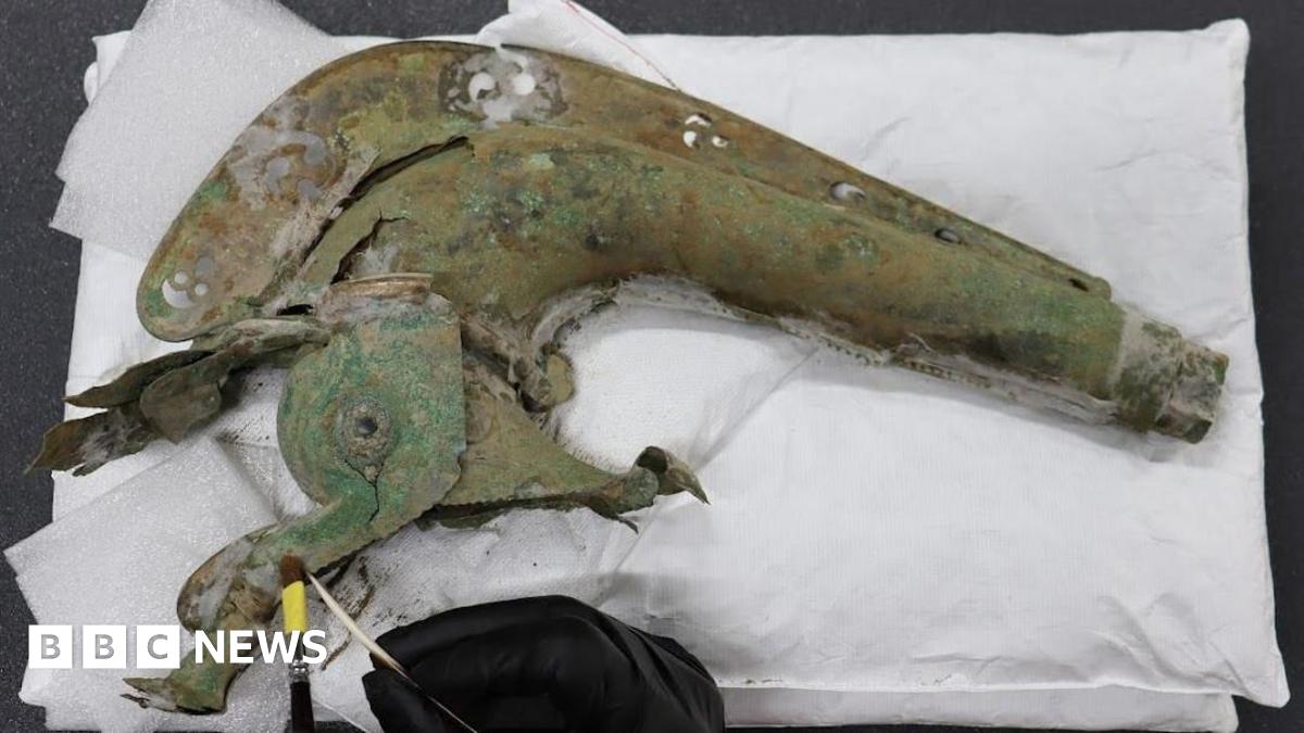 The head of an Iron Age carnyx emerging out of a block of sandy soil. It is lying on its side and half buried. It has the head of an animal, with a gaping mouth and crest.