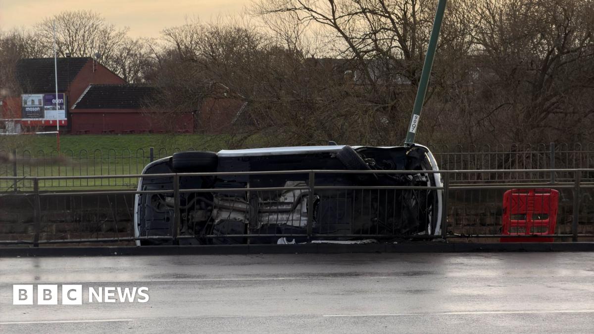 A car on its side, with its underside towards the camera, behind a pedestrian barrier next to a road, with parkland visible beyond