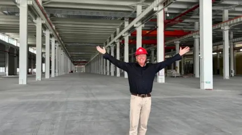 Craig Ledwards Craig Ledwards, smiles while he wears a red hard hat and stands with arms outstretched inside a large, empty industrial warehouse. The space features long rows of white support columns, a high ceiling with exposed beams and piping, and a wide concrete floor extending into the distance.