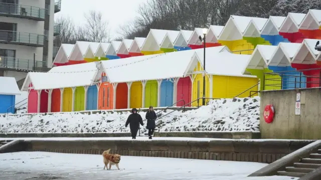 Dog walkers brave the wintry conditions in Scarborough as they walk past colourful beach huts