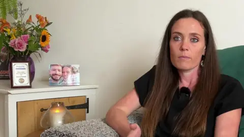 BBC Victoria Rodwell is sitting on a sofa at home. She has long brown hair and is wearing a black T-shirt. A candle is burning next to her and there is a vase of flowers on a cupboard, with a certificate from her 2022 Channel swim and her engagement picture.