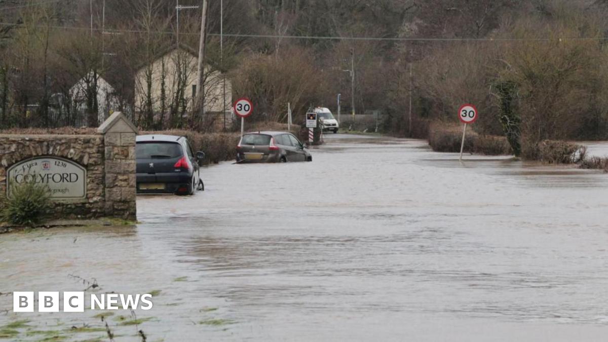 Two cars are submerged in floodwater on a road in Colyford, Devon. A sign with the village's name on it attached to a brick wall is in the foreground. A pair of 30mph signs are in the distance.