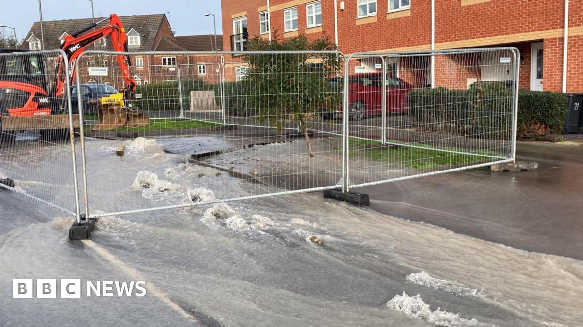 A street with houses down the far side and water running down the street with metal fencing closing off part of the street