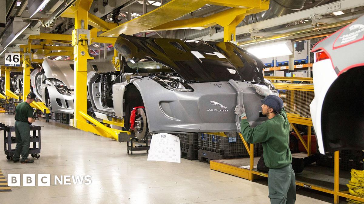 A man in a green jumper and blue cap closely inspects the grey body of a car which is suspended above a factory floor.