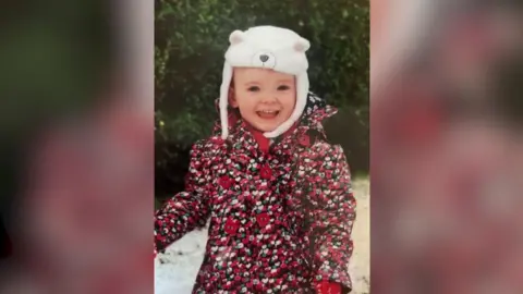 Walls Family Emily as a toddler, she is outside in the snow wearing a red floral coat and a white polar bear hat. She is smiling and has blonde hair. 
