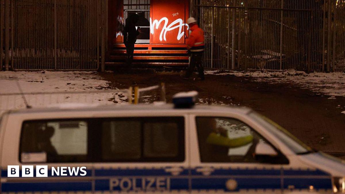 Police and emergency personnel wearing uniforms and one a helmet inspect a metal box with graffiti on it beside a fence in the dark. A police van is seen in the foreground and snow is on the ground.
