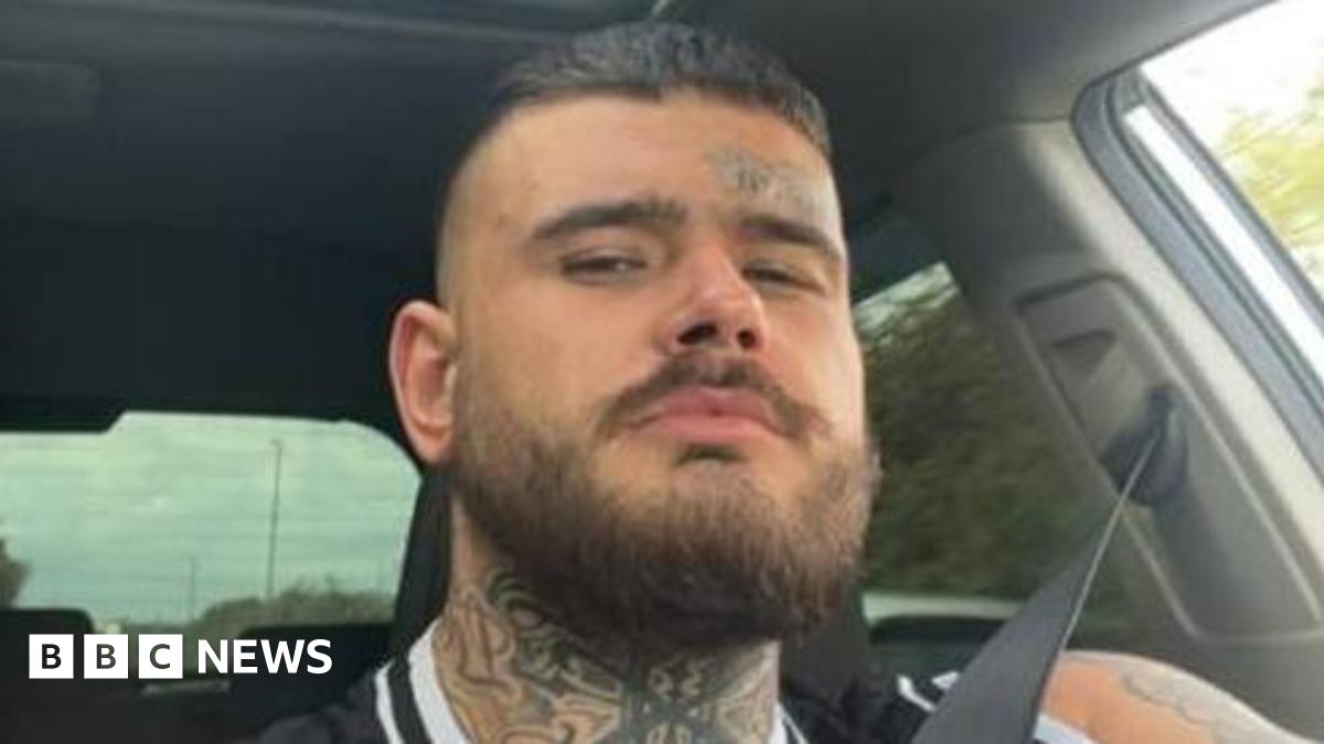 A selfie of a man sitting in a car. The trees in the background are blurred showing the car is moving. He is wearing a seatbelt and looking into the lens. He has a bear, a neck tattoo and a small tattoo above his eyebrow.