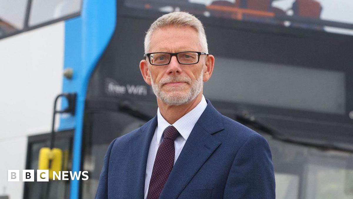 Darren Roe is in the middle. He has short grey hair and is wearing black glasses, a white shirt, burgundy tie and a blue blazer jacket. He is stood in front of a double decker bus.