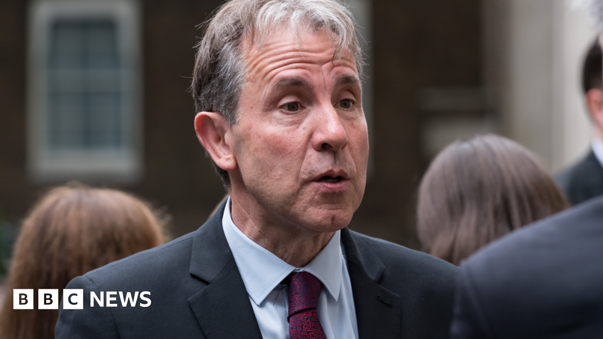 Dan Norris speaking to someone while wearing a navy suit and red and blue tie. He is stood outside with people behind him.