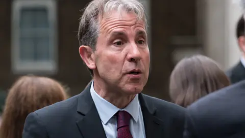 Getty Images Dan Norris speaking to someone while wearing a navy suit and red and blue tie. He is stood outside with people behind him.