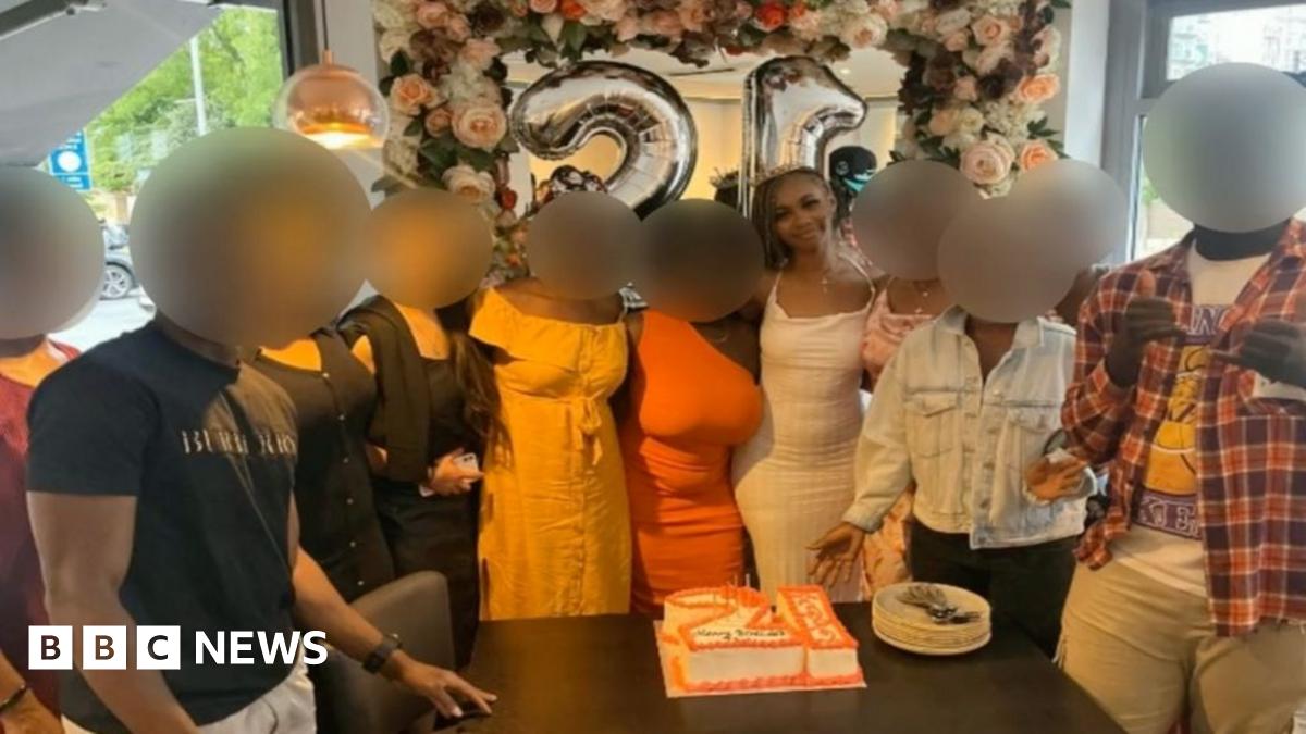 A group of 11 young people stand close together next to a cake in the shape of the number 21. All their faces are blurred apart from that of a young woman smiling in the middle.