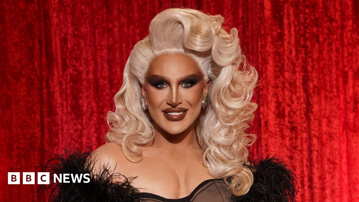 The Vivienne has curly blonde hair, is wearing a black off-the-shoulder dress and is smiling at the camera in front of a red velvet curtain.