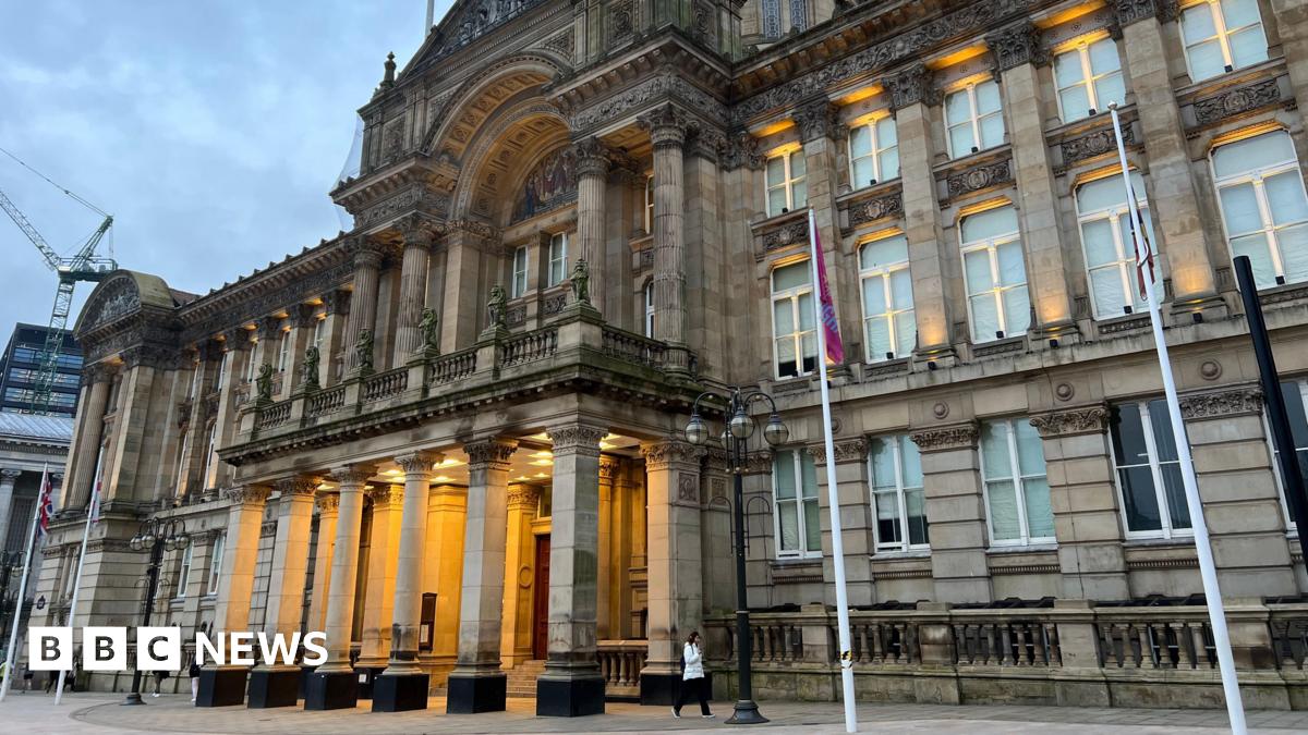 A large and imposing stone building with a columned portico at the front that is lit up, and a domed roof with four flag poles standing in front