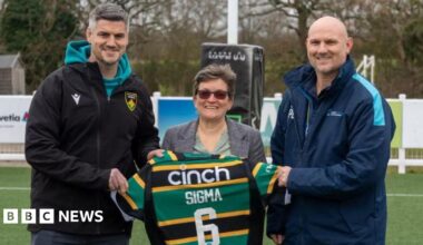 On the left, Mark Hopley has greying short hair and is wearing a turquoise hoodie underneath a black Northampton Saints coat. He is standing next to Stephanie Neill, who has short light brown hair and is wearing a tweed jacket. On the right is Peter Le Marquand, who is wearing a sports-style blue coat with a turquoise stripe on the shoulder. They are holding a rugby shirt, which says Sigma 6, and are standing on a pitch.