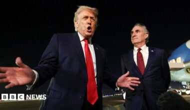 Donald Trump and Keir Starmer sit in a formal drawing room setting  on arm chairs with US and UK  flags behind them. Both have serious expressions and are looking out and not at each other.