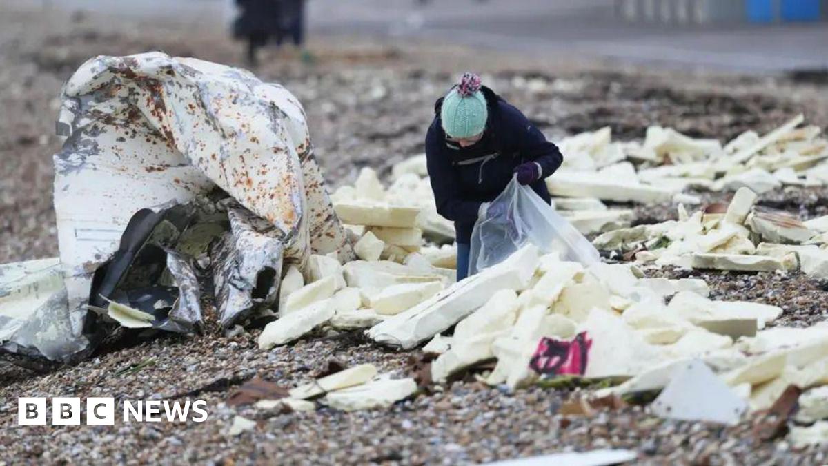A person putting white blocks of foam into a large, clear plastic bag on a pebble beach
