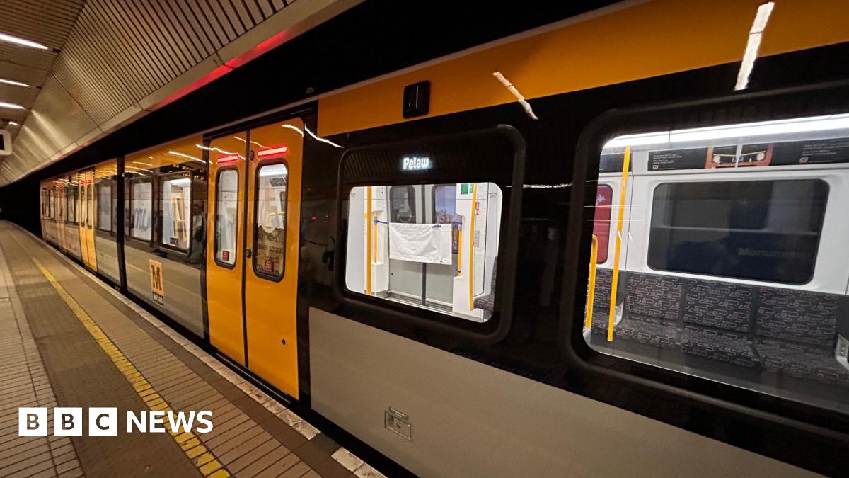A yellow, grey and black Metro train pulled into a station. The train is empty and its destination is Pelaw which is lit up on an LED display by the doors. The platform has a brown and yellow tiled floor.
