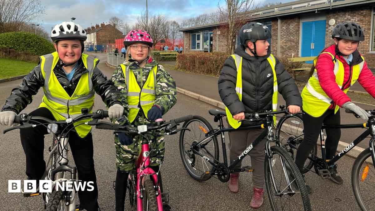 Four children are pictured on their bikes. They re all wearing high-vis jackets over their coats. A boy on the left is wearing a white helmet and smiling. A girl to his right is on a pink bike and wearing a pink and white spotted helmet. Two other boys are wearing black helmets.