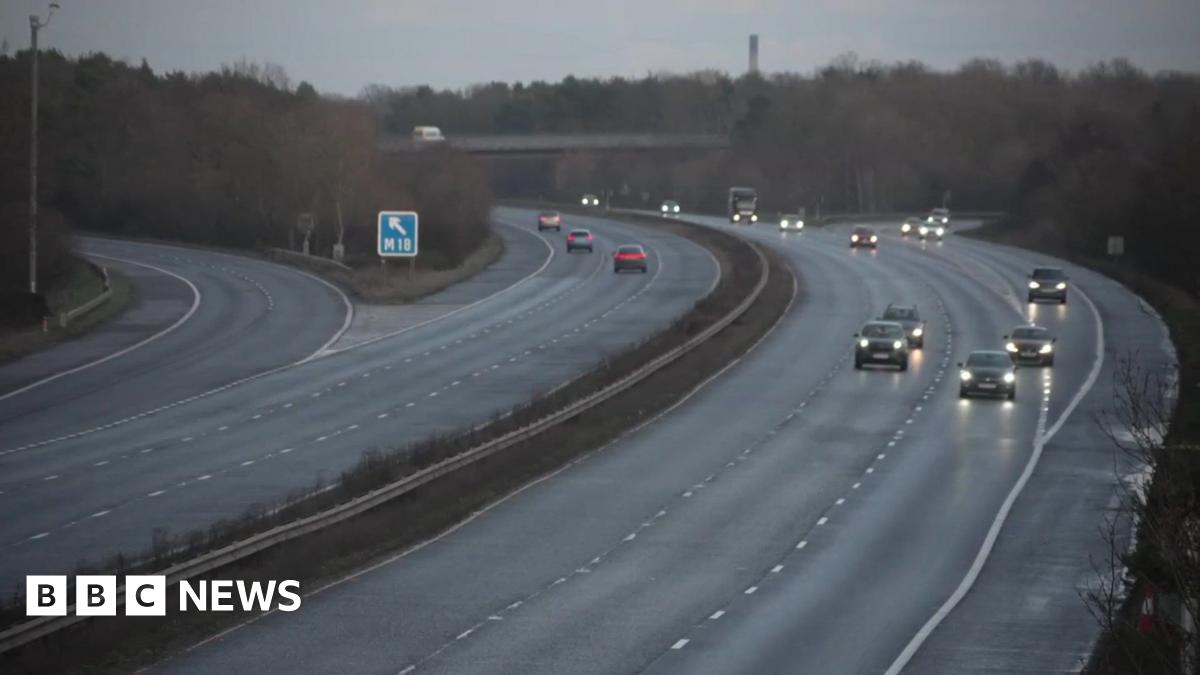 The M62 motorway with the east and westbound carriageways separated by a strip of grass and metal central reservation barrier. A number of vehicles can be seen on both sides of the road and have their headlights turned on. Trees and a bridge are visible in the distance along with a blue road sign.