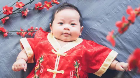 Getty Images A baby lying down on a patterned grey cloth while dressed in a red traditional Chinese outfit with gold linings. Some red flowers surround him. 